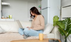 Вид збоку жінка друкує на нетбуці, сидячи на дивані в будинку — стокове фото