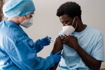Жіночий лікар в захисній формі і латексні рукавички вакцинують чоловічого афроамериканського пацієнта в клініці під час спалаху коронавірусу. — стокове фото