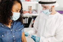 Побочный обзор врача-мужчины в защитной форме, латексных перчатках и защитном щите для прививки афроамериканской пациентки в клинике во время вспышки коронавируса — стоковое фото