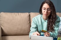 Jovem mulher feliz usando seu cartão de crédito amarelo para fazer compras on-line com tablet, sentado no sofá em casa — Fotografia de Stock