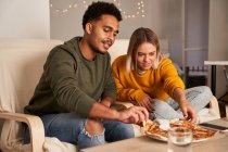 Multiethnisches Paar sitzt in Sesseln und isst leckere Pizza, während es das Wochenende zu Hause zusammen genießt — Stockfoto