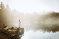 Жінка стояла в білій сукні на скелі, дивлячись на озеро в туманний день — стокове фото