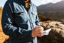 Вид збоку врожаю невпізнаваний дослідник чоловічої статі, що стоїть на пагорбі в горах і пише в блокноті під час відпустки — стокове фото