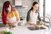 Jóvenes novias lesbianas cortando pepino mientras preparan almuerzo vegano en la mesa con vasos de bebidas alcohólicas en casa - foto de stock
