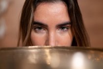 Crop young female covering mouth with Himalayan bowl while looking at camera on blurred background — Stock Photo