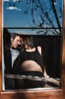 A través de la ventana de la mujer embarazada y el hombre contento tiernamente tocando las narices en el día soleado en casa - foto de stock