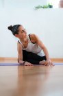 Nivel del suelo de la mujer de mediana edad estirando las piernas y la espalda mientras practica yoga en la estera y mirando hacia otro lado en la habitación - foto de stock