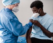 Vista lateral del médico femenino en uniforme protector y guantes de látex que desinfectan el brazo con algodón con alcohol para vacunar al paciente afroamericano masculino en la clínica durante el brote de coronavirus - foto de stock