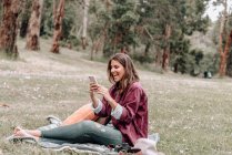 Mujer alegre en sombrero sentado en manta en el prado en el bosque y la navegación del teléfono móvil mientras disfruta de un picnic en Australia - foto de stock