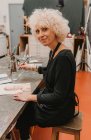 Вид збоку весела жінка-художник сидить за столом і малює акварелями під час роботи в художній студії і дивлячись на камеру — стокове фото