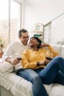 Content multiracial couple chilling on sofa at home with red wine in glasses while enjoying weekend at home and looking at each other — Stock Photo