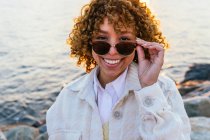 Femme afro-américaine joyeuse sur des lunettes de soleil élégantes debout sur le bord de la mer et profiter de la liberté au coucher du soleil en regardant la caméra — Photo de stock