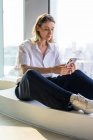 Самотня неемоційна молода жінка сидить у порожньому офісі з великим вікном, що переглядає мобільний телефон — стокове фото