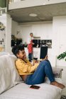 Fröhliche Afroamerikanerin liest interessantes Buch auf Sofa vor dem Hintergrund eines Mannes, der in der Küche kocht — Stockfoto