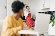 Homem de avental falando no smartphone na cozinha e mulher negra navegando laptop em casa — Fotografia de Stock