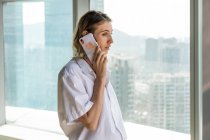 Молода вдумлива бізнес-леді, що стоїть в офісі з великими вікнами, має телефонний дзвінок на мобільний телефон — стокове фото