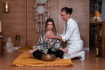 Zen-Lehrer in weißer Kleidung berührt Brust einer Frau mit gekreuzten Beinen während Yoga-Praxis in der Nähe des Schüsselgong — Stockfoto