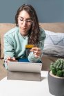 Young happy woman using her yellow credit card to shop online with tablet, sitting on the sofa at home — Stock Photo