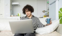 Jovem hipster macho com cabelos cacheados navegando na internet no netbook enquanto descansa no sofá na sala da casa — Fotografia de Stock