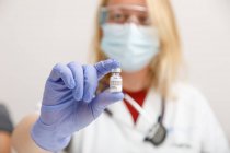 Female medic in protective face shield mask and latex gloves with vial of coronavirus vaccine showing to camera while standing in hospital room — Stock Photo