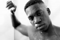 Bianco e nero del giovane ragazzo nero senza emozioni che si fa la doccia in bagno leggero e guarda la fotocamera e l'acqua sul viso — Foto stock