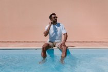 Серьезный мужчина-фрилансер, сидящий у бассейна с ногами в воде и разговаривающий на мобильном телефоне во время удаленной работы летом — стоковое фото
