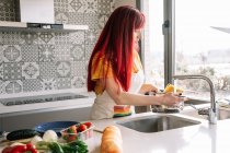 Молодая гомосексуальная женщина наливает воду из крана в кастрюлю с сырой пастой против разнообразных овощей в доме — стоковое фото