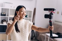 Smiling ethnic female vlogger recording video on photo camera while making hand gestures standing in living room — Stock Photo