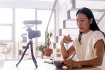Молода етнічна жінка відеоблогер з блокнотом, що сидить за столом з фотоапаратом на тринозі на кухні — стокове фото