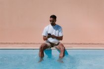 Freelance masculin sérieux assis au bord de la piscine avec les jambes dans l'eau et parlant sur un téléphone portable pendant le travail à distance en été — Photo de stock