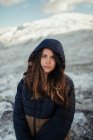 Young gentle female traveler in hood looking at camera against snowy mountains in winter on sunny day — Stock Photo