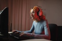 Vista lateral do gamer fêmea encantado em fones de ouvido jogando videogame enquanto sentado à mesa em casa — Fotografia de Stock