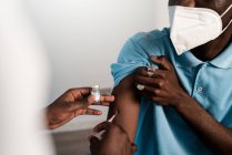 Médecin noir cultivé en uniforme de protection et gants en latex vaccinant un patient afro-américain masculin méconnaissable en clinique pendant l'épidémie de coronavirus — Photo de stock