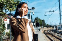 Desde abajo vista lateral de contenido viajero femenino étnico de pie en la plataforma en la estación de tren y hablando en el teléfono inteligente - foto de stock