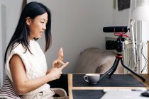 Усміхнена етнічна жінка блогер з чашкою гарячого напою говорить під час запису відео на фотокамеру вдома — стокове фото