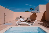 Vista lateral del freelancer masculino tumbado en la tumbona junto a la piscina y navegando por Internet en el portátil durante el teletrabajo en verano en un día soleado - foto de stock