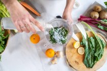 From above crop unrecognizable adult female pouring vegetarian milk into bowl with chard leaves and orange slices while preparing smoothie at home — Stock Photo