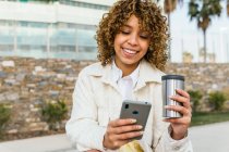 Mulher afro-americana positiva com mensagens de café takeaway nas mídias sociais via smartphone na rua da cidade — Fotografia de Stock