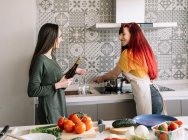 Content young homosexual girlfriends with bottle of wine speaking while looking at each other between stove and fresh vegetables — Stock Photo