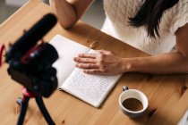 Cropped unrecognizable female vlogger with notebook sitting at table with photo camera on tripod in kitchen — Stock Photo