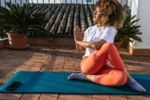 Donna etnica in abbigliamento sportivo seduta con gambe incrociate e mani Namaste mentre pratica yoga e distoglie lo sguardo sul tetto della casa — Foto stock