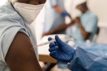 Médico en uniforme protector y guantes de látex vacunando a un paciente afroamericano irreconocible en la clínica durante el brote de coronavirus - foto de stock