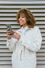 Optimistic African American female with Afro hairstyle browsing on smartphone while standing against metal wall in urban area in city — Stock Photo