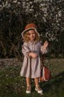 Восхитительная маленькая девочка в платье, стоящее в весеннем саду с цветущими цветами на деревьях и смотрящая в камеру, наслаждаясь солнечным днем — стоковое фото