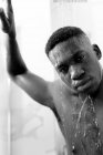 Чорно-білий без емоцій молодий чорний хлопець приймає душ у світлій ванній кімнаті і дивиться на камеру і воду на обличчі — стокове фото