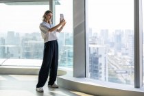 Молода жінка стоїть у порожньому офісі з великими вікнами, приймаючи селфі на мобільний телефон — стокове фото
