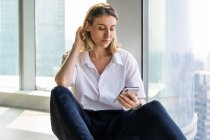Einsame, emotionslose junge Frau sitzt in leerem Büro mit großem Fenster und surft auf dem Handy — Stockfoto