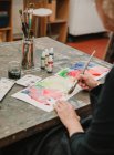 Crop unrecognizable female artist painting with watercolors on paper while sitting at table in art studio — Stock Photo
