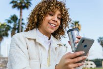 De baixo positivo afro-americano fêmea com cabelo encaracolado segurando takeaway mensagens de café nas mídias sociais via smartphone na rua da cidade — Fotografia de Stock