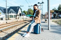 Smiling Asian female entrepreneur reading documents while sitting on suitcase at railway station and waiting for departure — Stock Photo